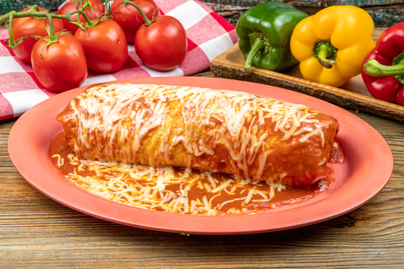A wet burrito on a coral oval plate, covered in red salsa and melted cheese, with vine tomatoes and bell peppers behind it on a rustic wood table.