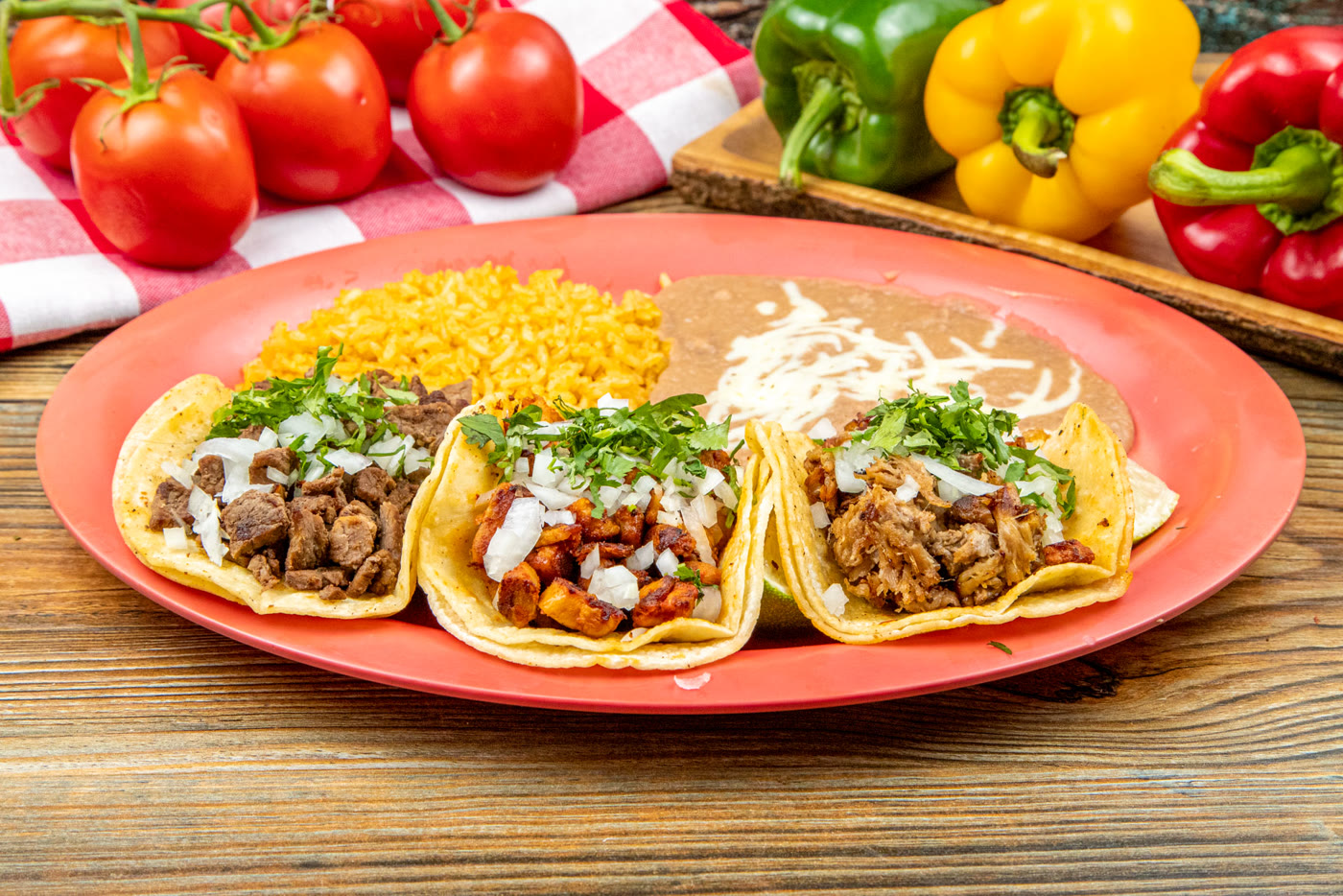 Three street tacos on a coral plate, one carne asada, one al pastor and one carnitas, each topped with chopped onion and cilantro, with yellow rice and refried beans alongside.