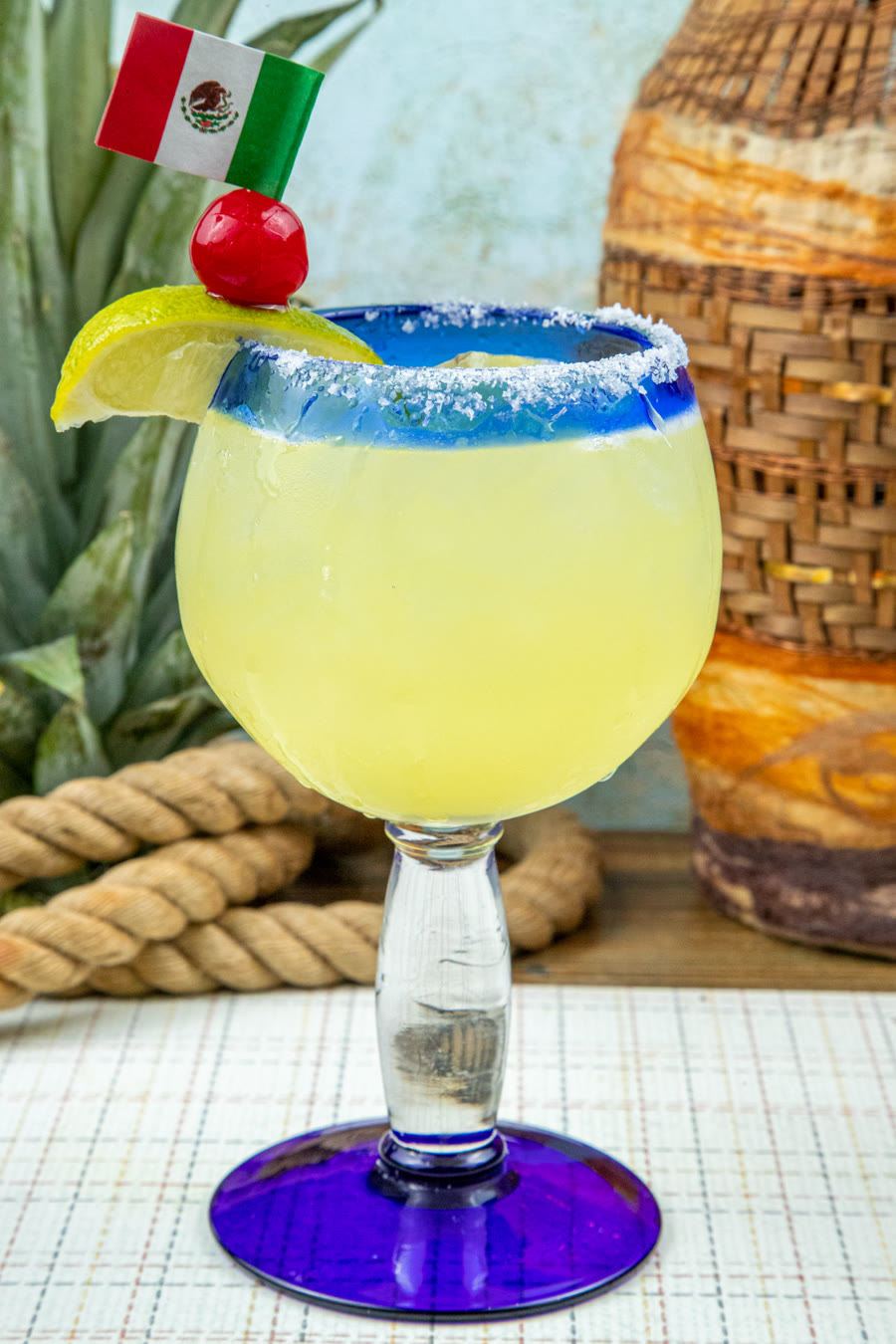 A classic margarita in a blue rimmed glass with a salted rim, garnished with a cherry and a lemon wedge on a small Mexican flag pick, with a pineapple and a woven basket behind it.
